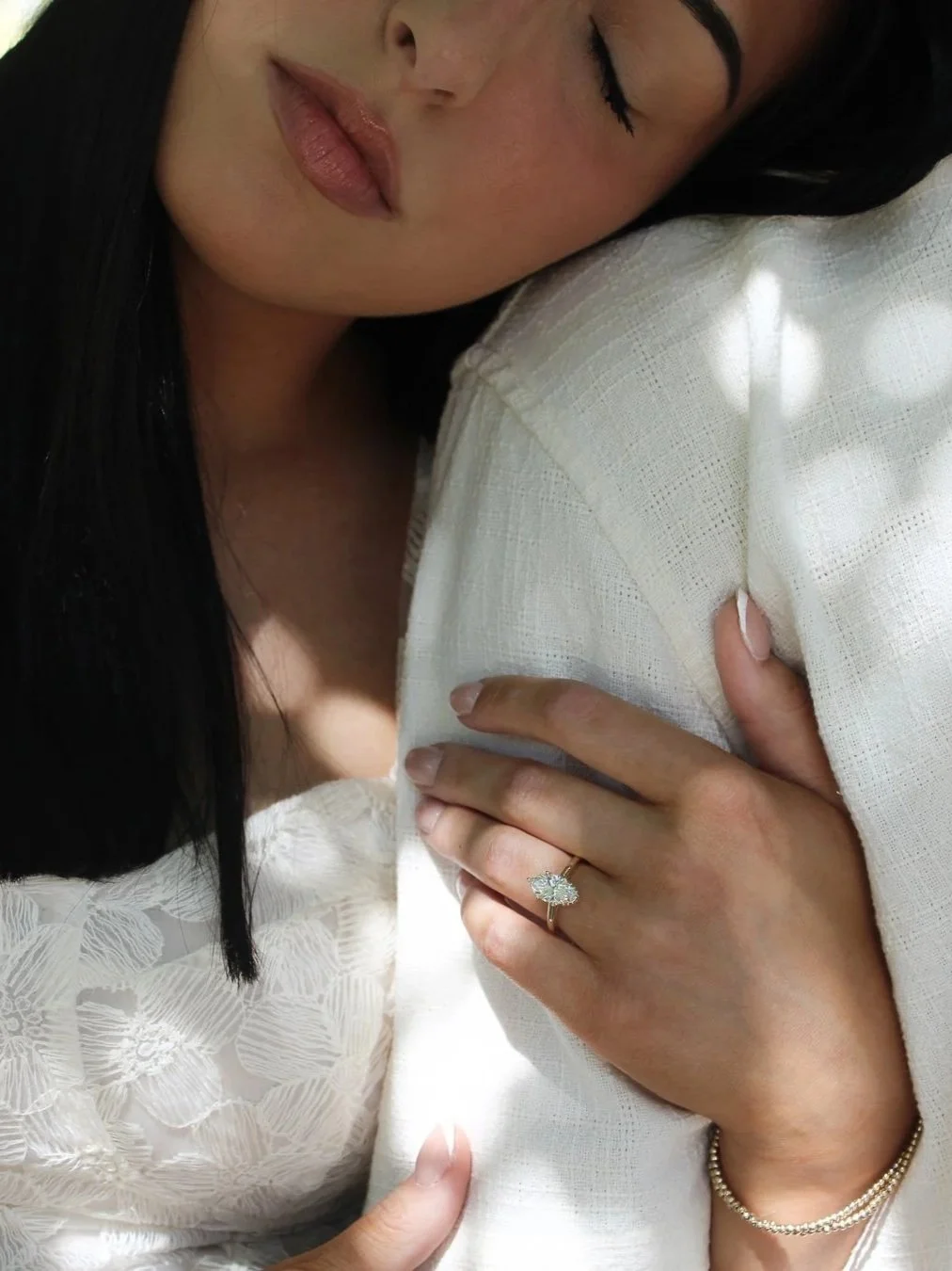 Woman with black hair wearing a white dress,  leaning her head on her fiancé's shoulder with her eyes closed.