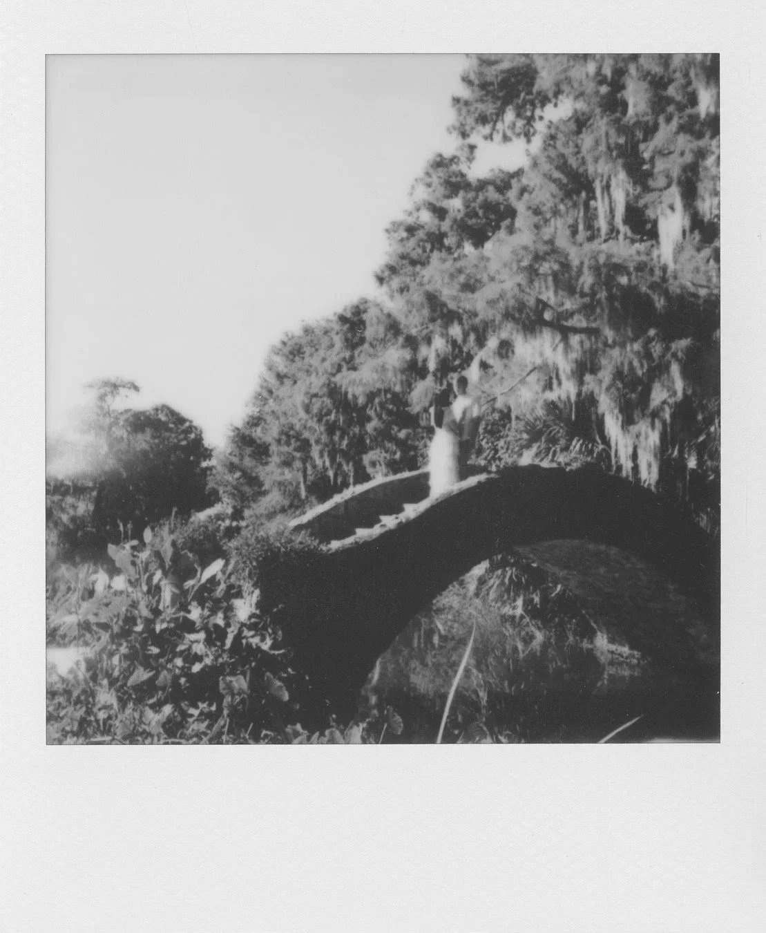 Black and white Polaroid of woman and man wearing white standing on bridge surrounded by nature.