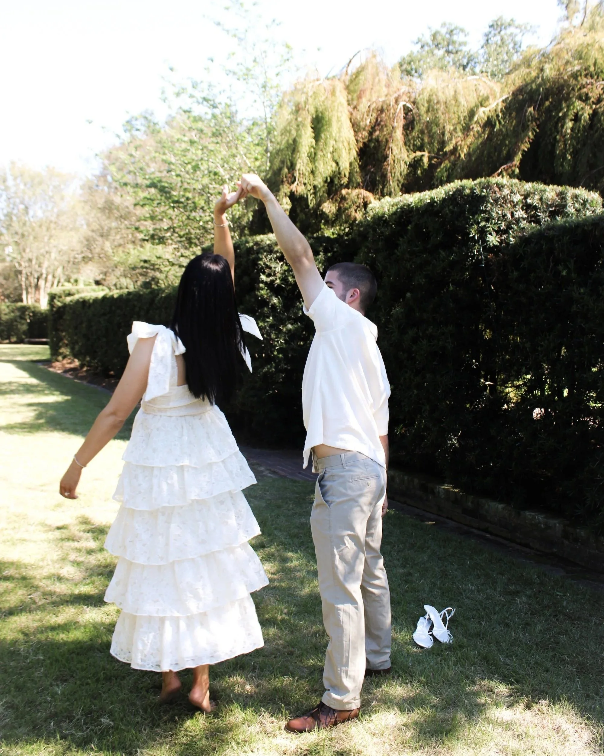 A couple holding hands and dancing outdoors on a sunny day, with the woman wearing a white ruffled dress and the man in beige pants and a white shirt, standing on a grassy area with tall bushes in the background.