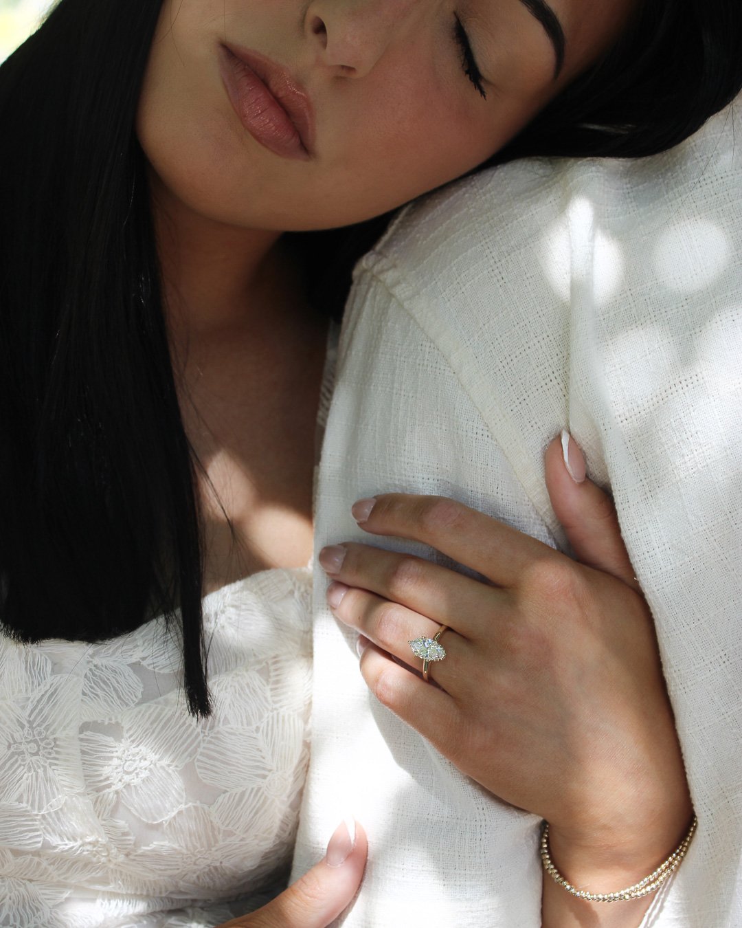 A woman with dark hair, wearing a white lace dress, is resting her face on her fiancé's shoulder. She has her eyes closed, and her hand wearing her engagement ring is gently placed on the shoulder. She is also wearing a gold bracelet.
