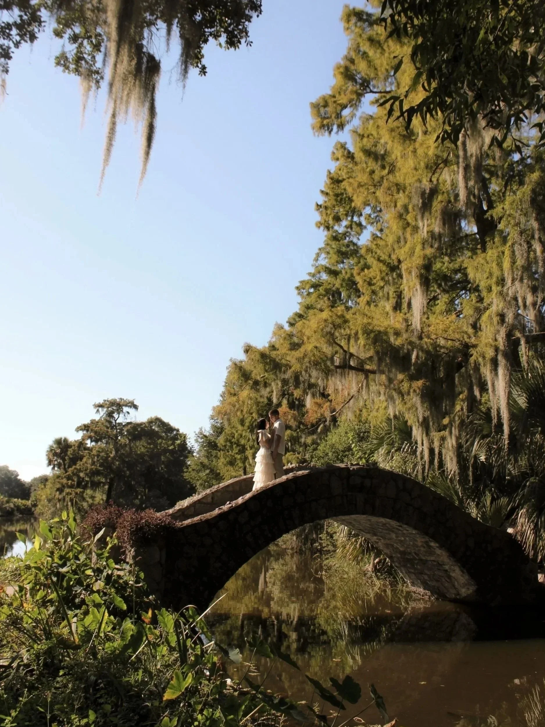 Woman and man wearing white standing on bridge with greenery surrounding them.