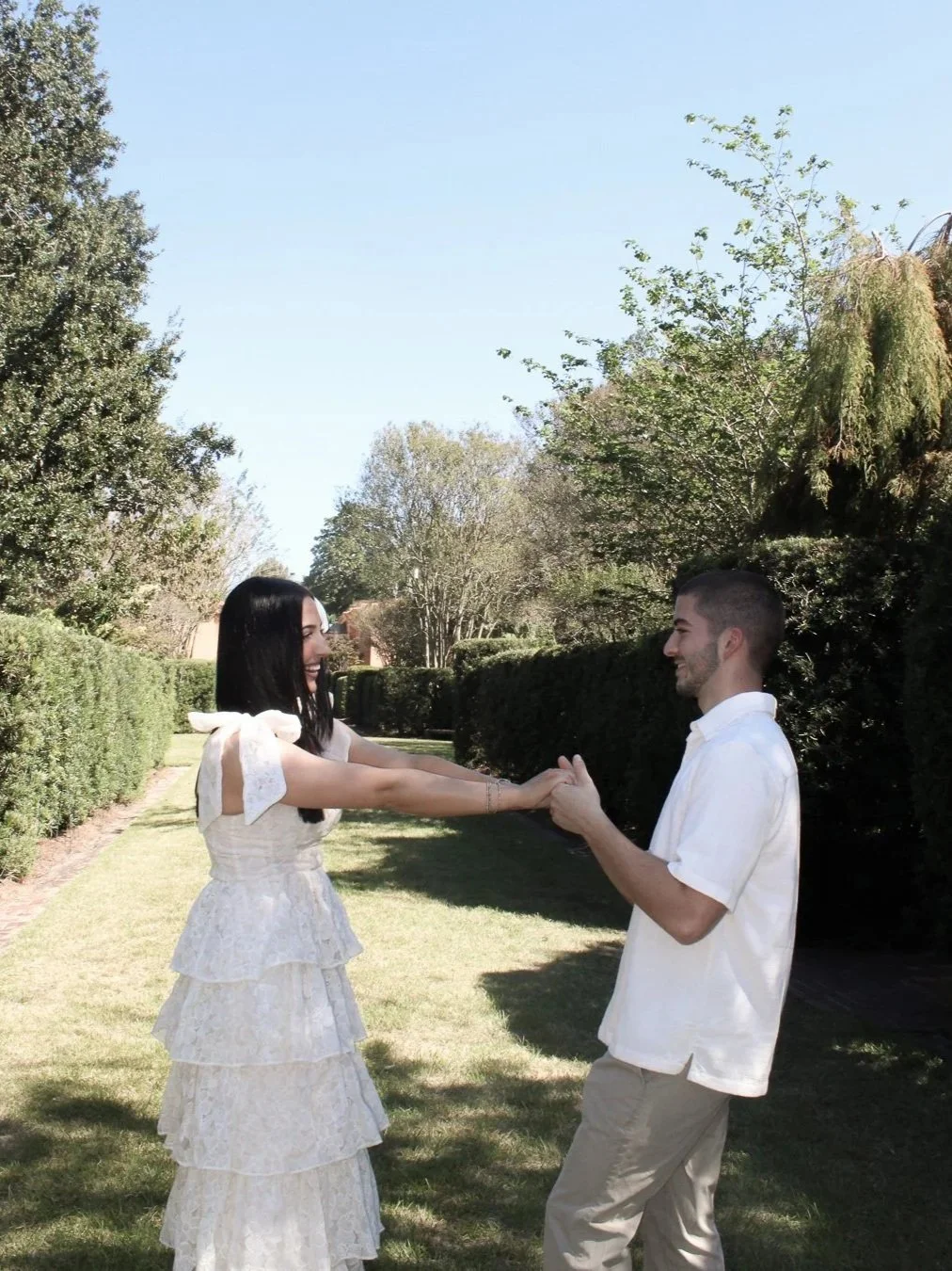 Woman and man dancing in garden, both wearing white outfits and smiling. Greenery surrounds them.