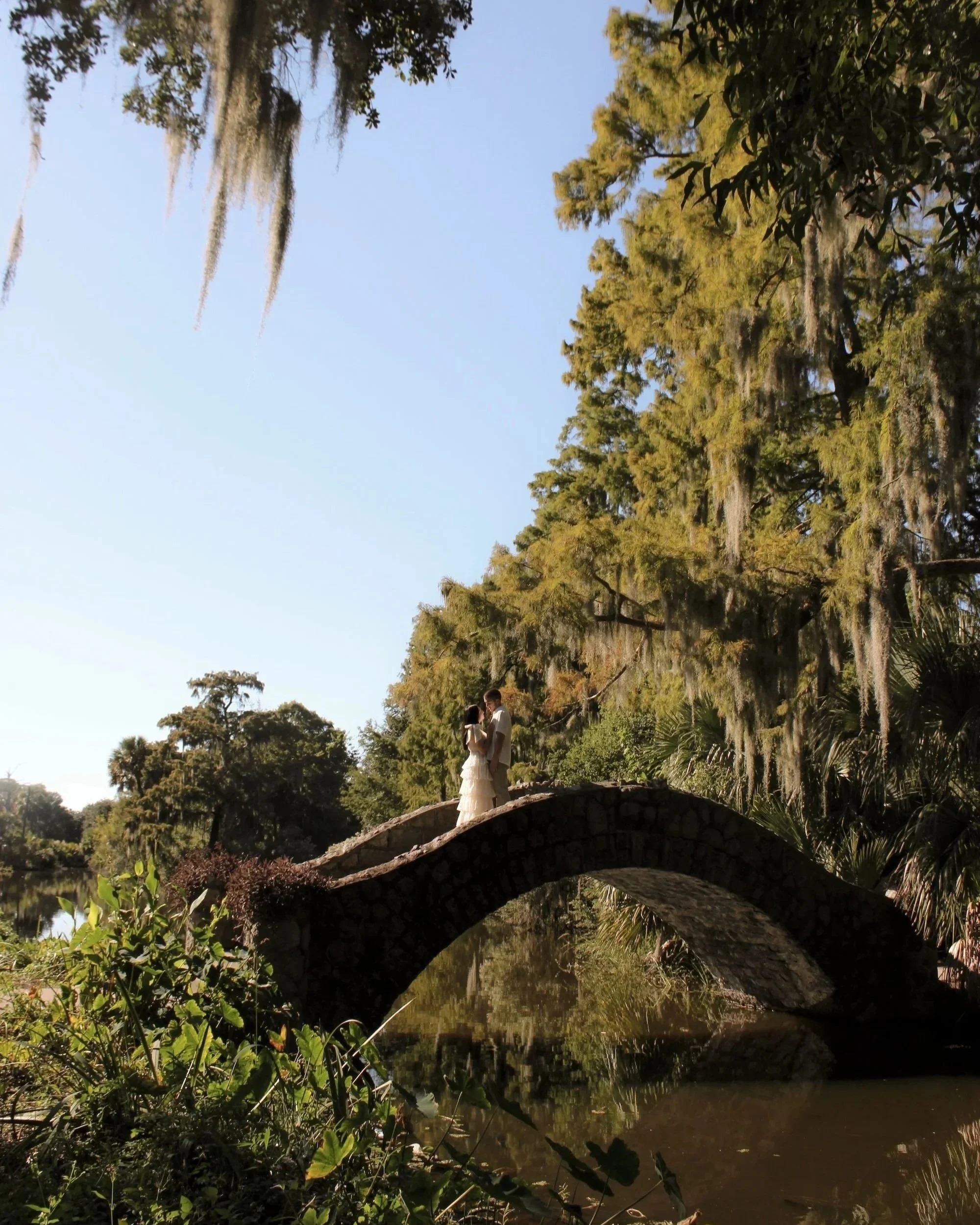 A couple in white clothing standing on a stone bridge over a calm river, surrounded by lush green trees with moss hanging from branches on a clear day.