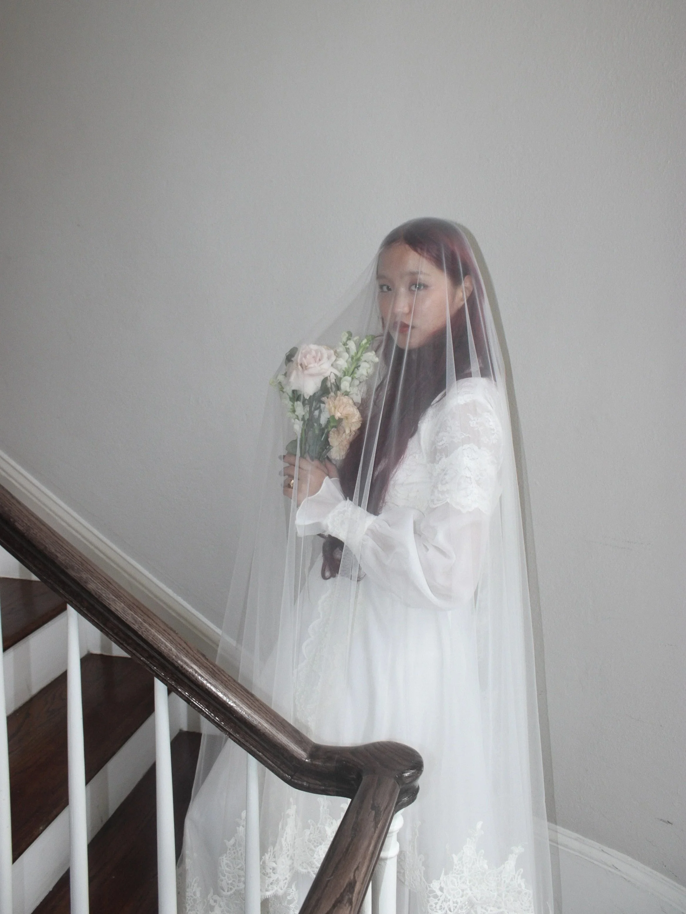 Woman with long hair is looking at camera and wearing a long sleeve white lace wedding dress. She is holding her bouquet and standing on wooden stairs.