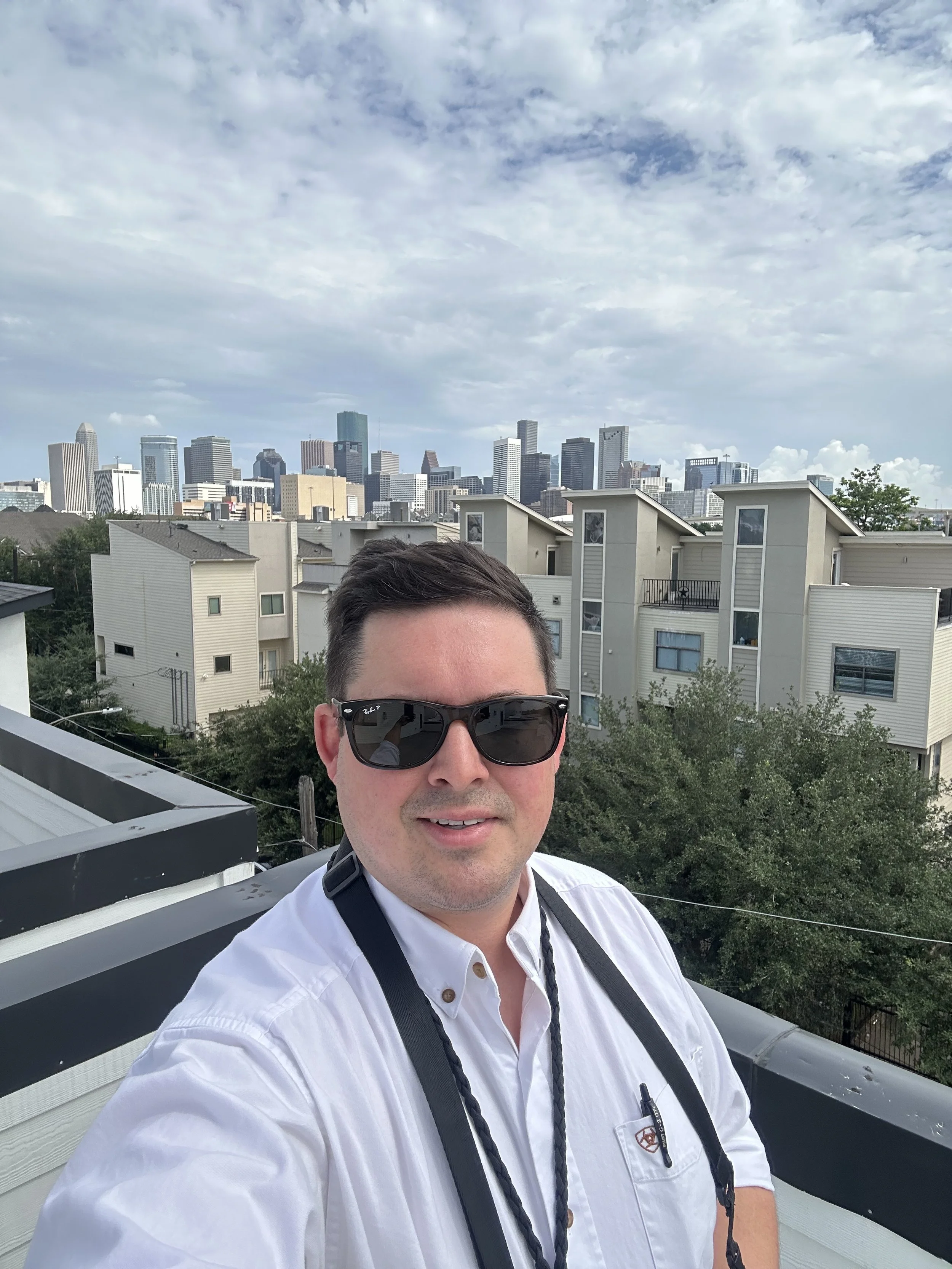 A man wearing sunglasses and a white shirt with a camera strap around his neck taking a selfie on a balcony with a city skyline in the background.