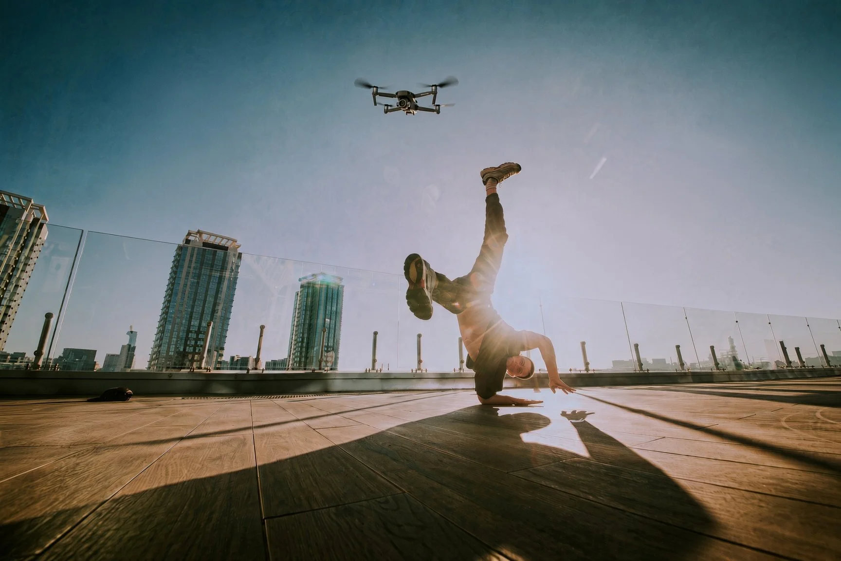 A person in a handstand yoga pose on a rooftop with city buildings in the background, drone flying overhead, and sunlight creating lens flare.