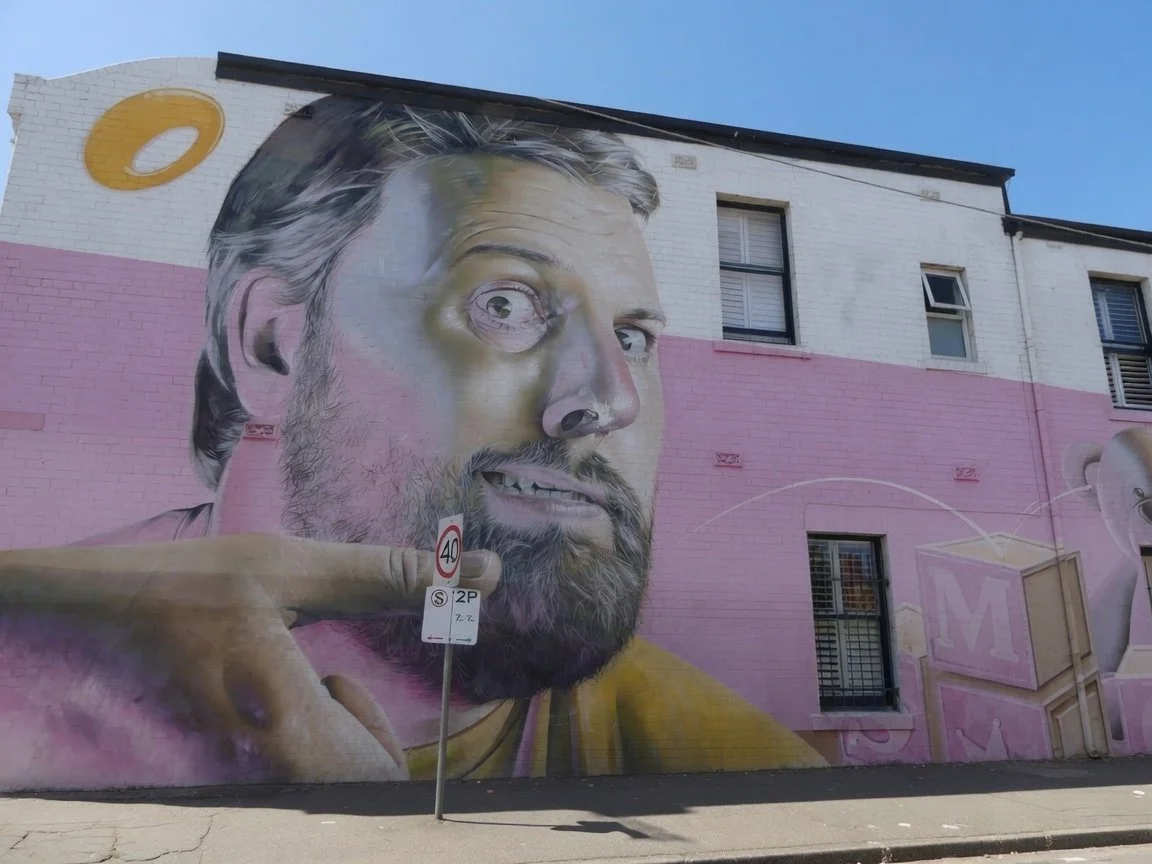 A large mural of a man with a beard and gray hair on the side of a building, with a pink background and a building in the top right corner. The man is holding his hand near his chin, with a street sign showing a 40 km/h speed limit in front of the mural.