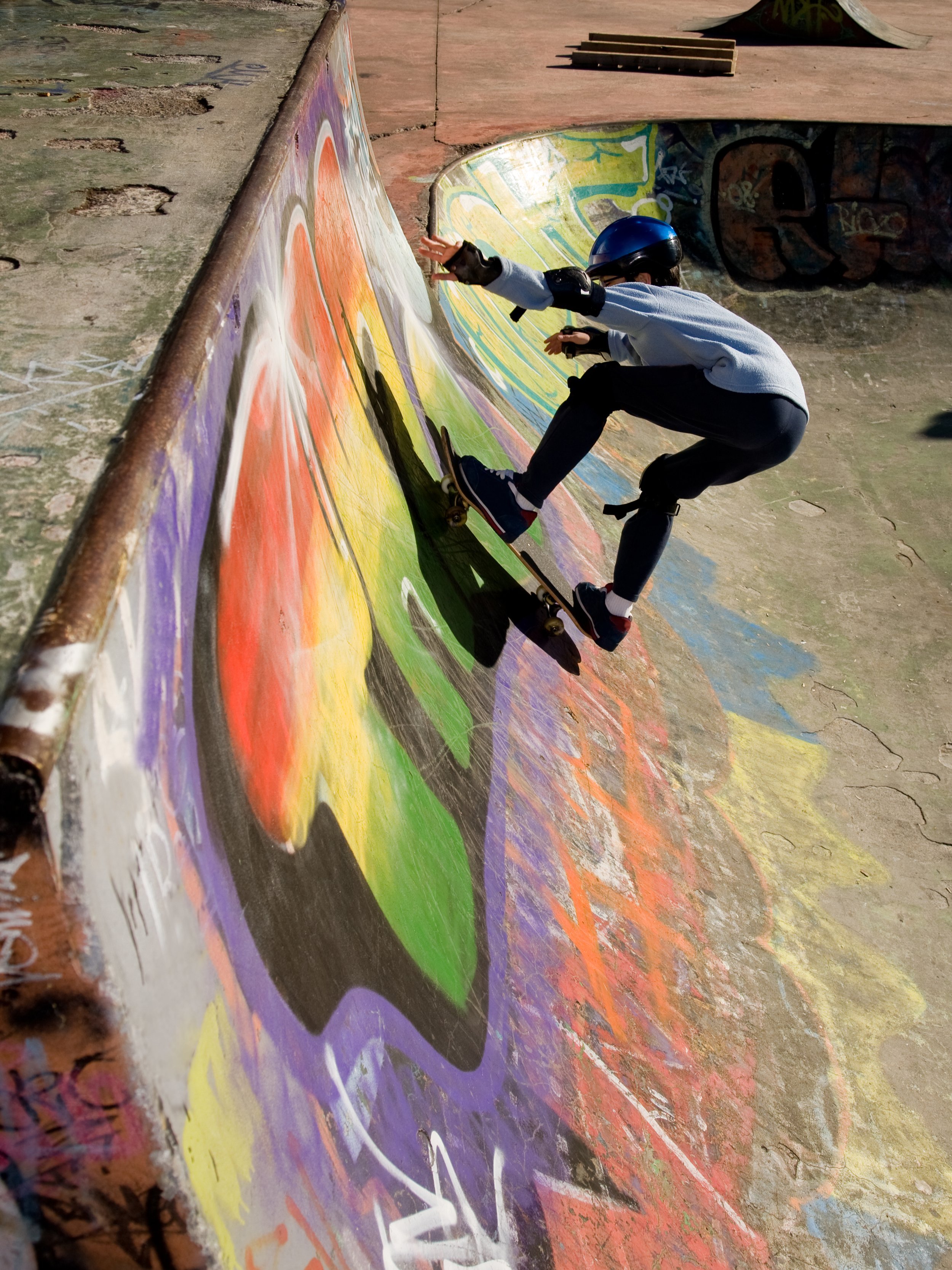 A person in a helmet and gray sweatshirt doing a skateboarding trick on a colorful graffiti-covered skate park ramp.