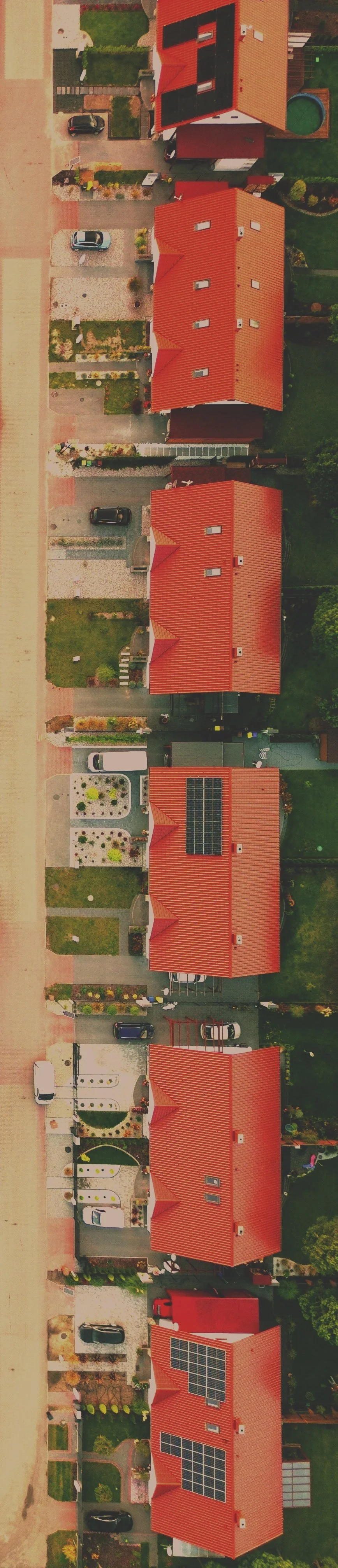 Aerial view of multiple houses with red roofs, some with solar panels, in a suburban neighborhood, with driveways, cars, and green lawns.