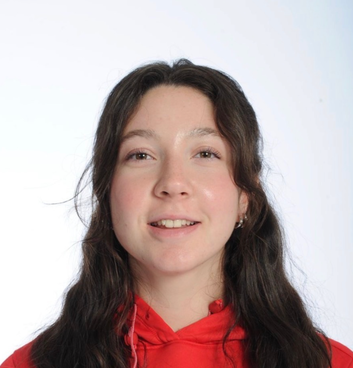 A young woman with long wavy brown hair wearing a red hoodie and smiling at the camera against a plain white background.