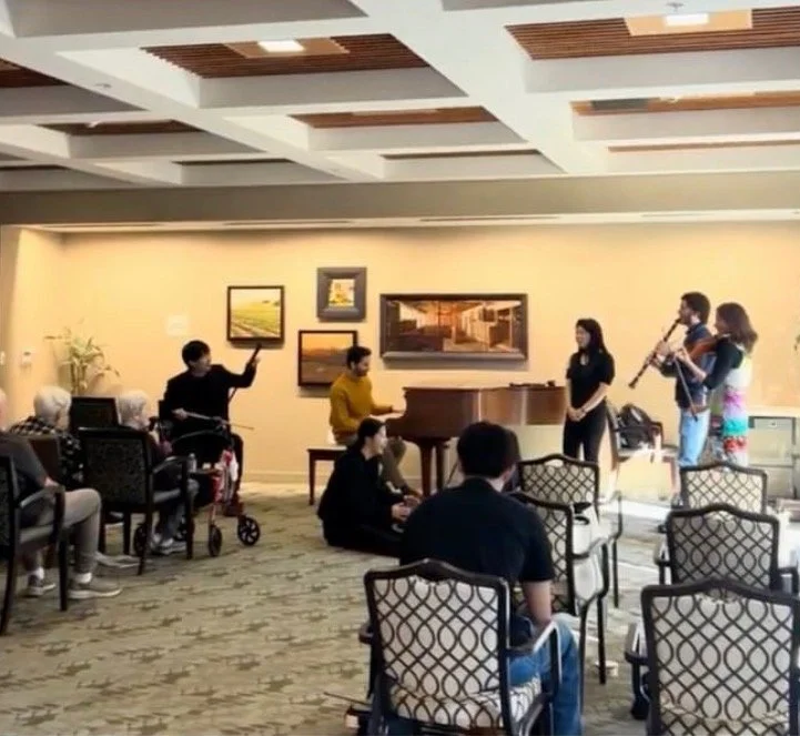 Group of people performing in a room with chairs, a piano, and artwork on the wall, while an audience listens.