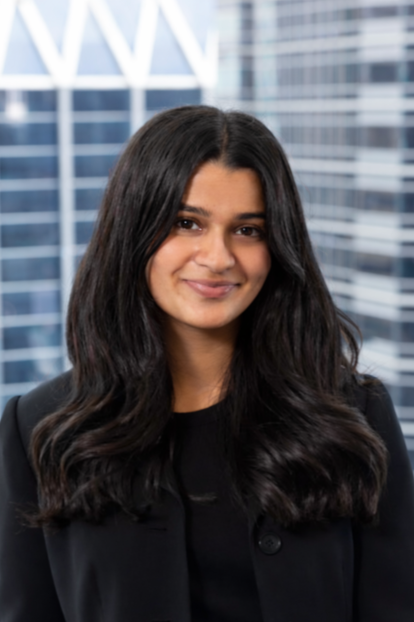 Aliza Vriji - A woman with long dark hair smiling, wearing a black blazer, standing in front of tall glass office buildings.