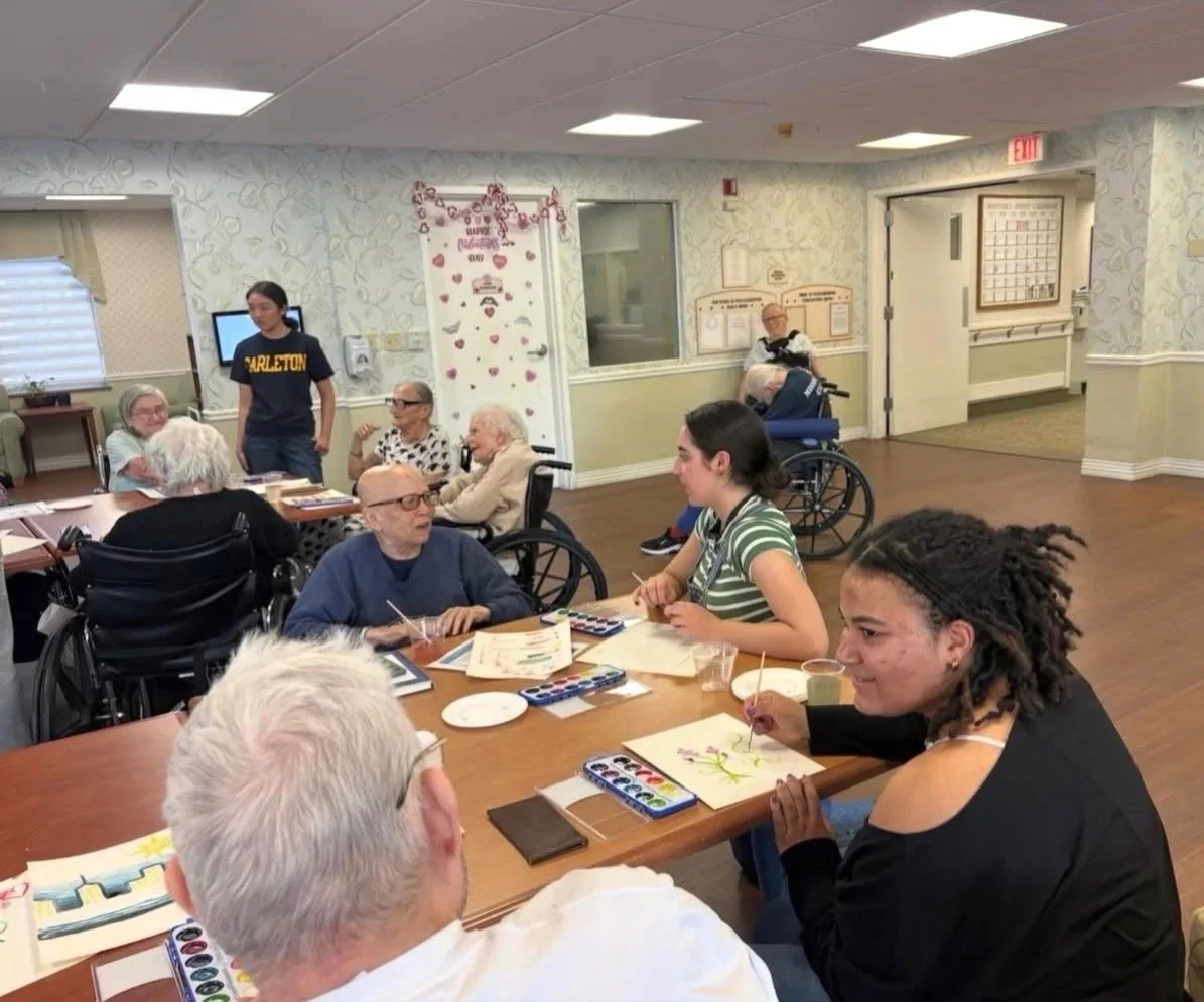Group of elderly and young people participating in an arts and crafts activity in a communal room, with some in wheelchairs and some standing or sitting at tables, painting and drawing.