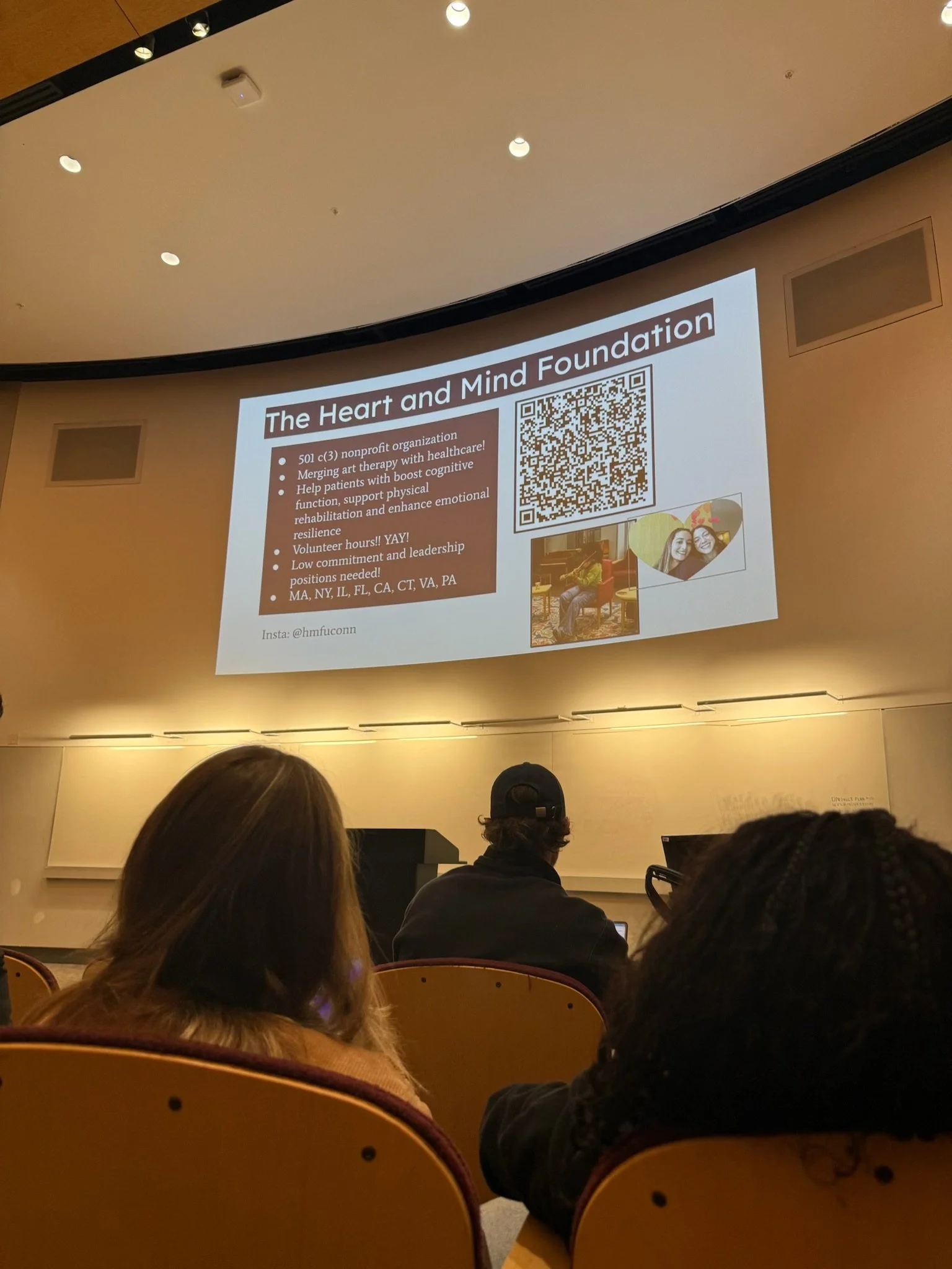 A presentation slide titled 'The Heart and Mind Foundation' is projected on a large screen in a conference room. The slide contains a QR code, some text about the organization, and a few images of people, including two women smiling and a person play