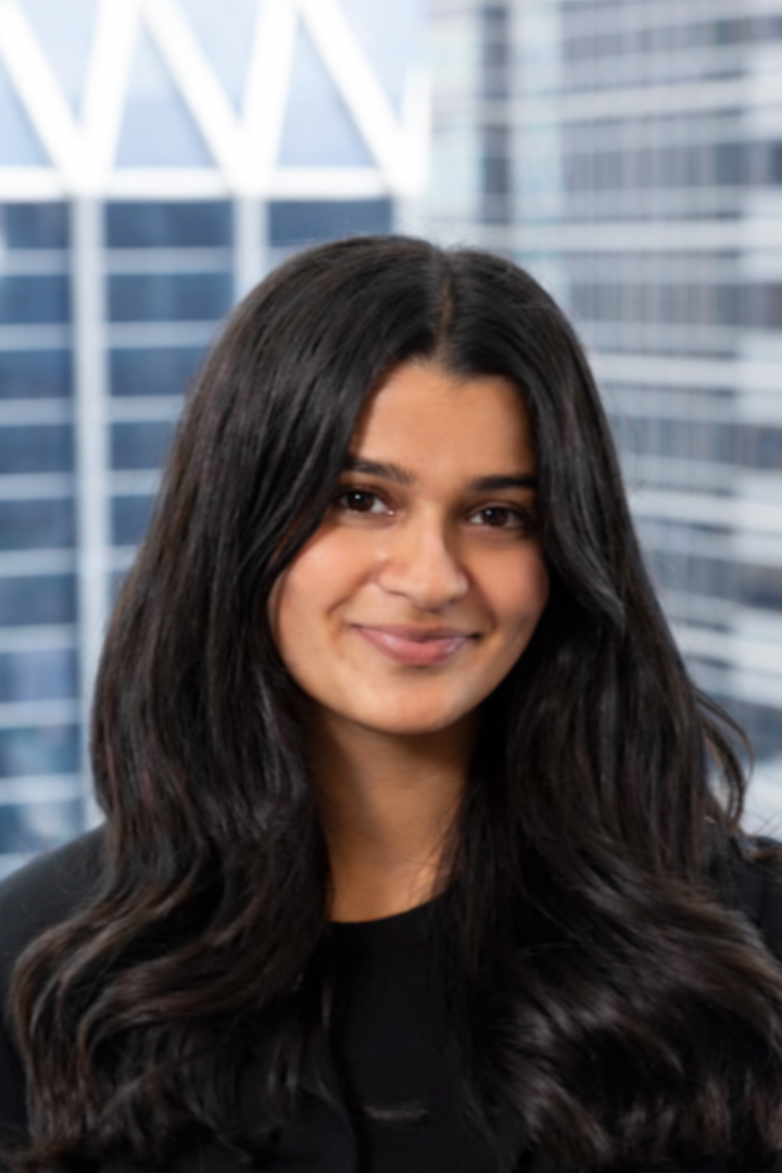 Aliza Vriji - A woman with long dark hair smiling, wearing a black blazer, standing in front of tall glass office buildings.