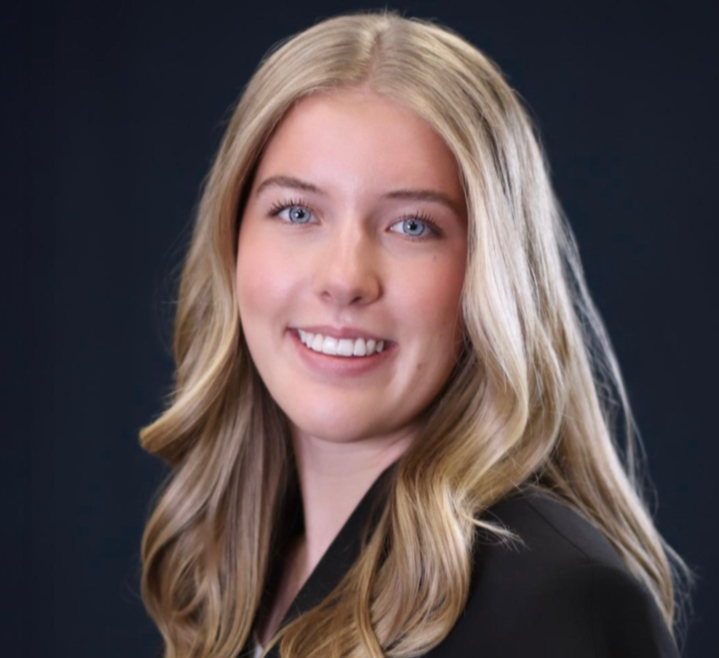 A young woman with long, blonde hair and blue eyes, smiling, dressed in a black blazer, against a dark background.