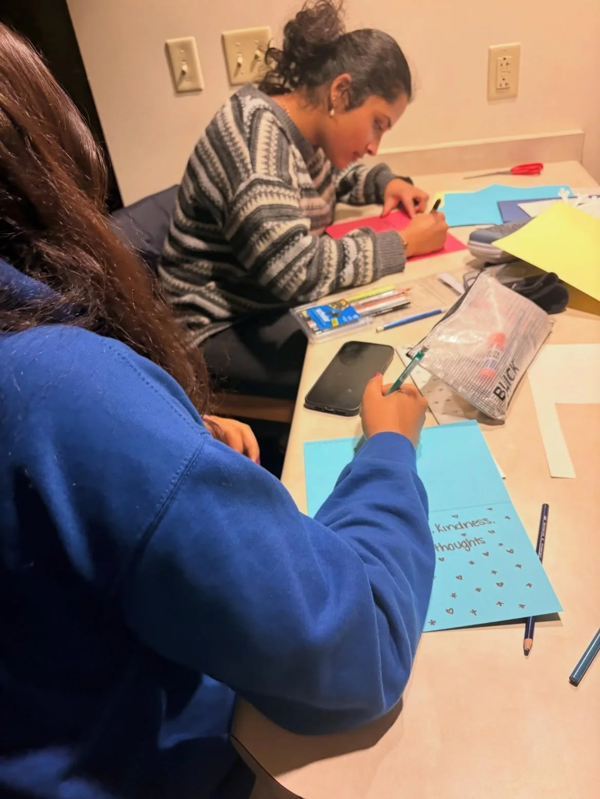 Two women sitting at a table working on papers, one writing on a blue sheet that says 'Kindness thoughts,' and the other writing on a red sheet with various art supplies scattered on the table.