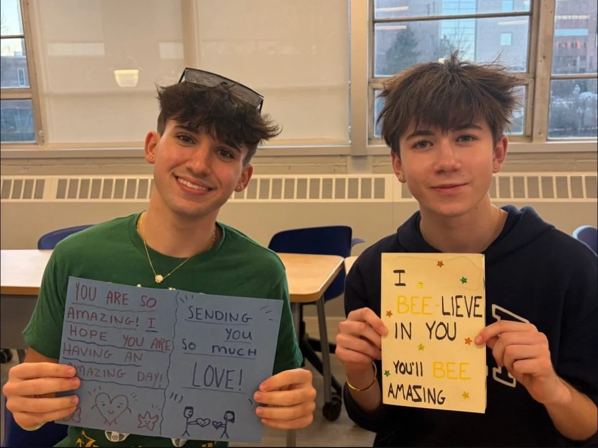 Two young men holding handmade signs with positive messages, sitting in a room with large windows in the background.
