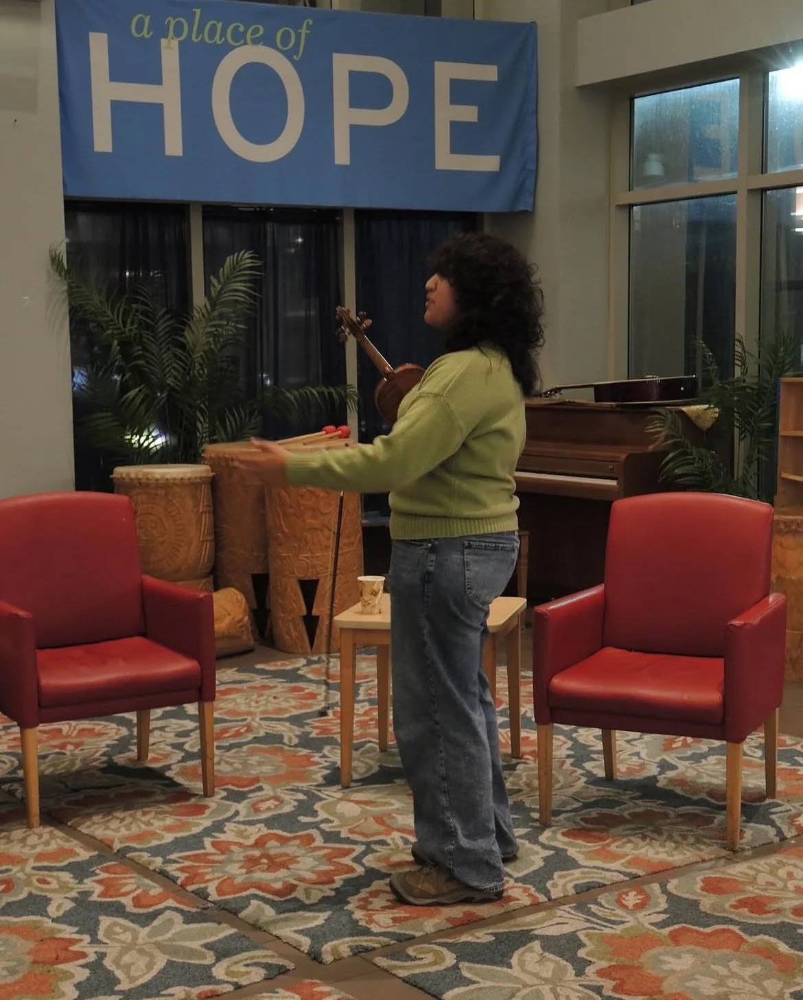 A woman with curly hair playing a musical instrument, possibly a violin, in a cozy room with red chairs, a potted plant, a patterned rug, and a large banner that reads 'a place of HOPE' in the background.
