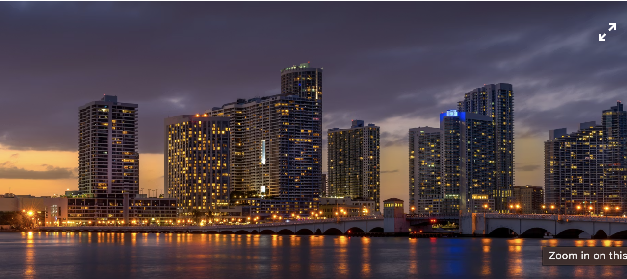 A city skyline during sunset with tall skyscrapers, highways, and roads in the foreground, and a partly cloudy sky.
