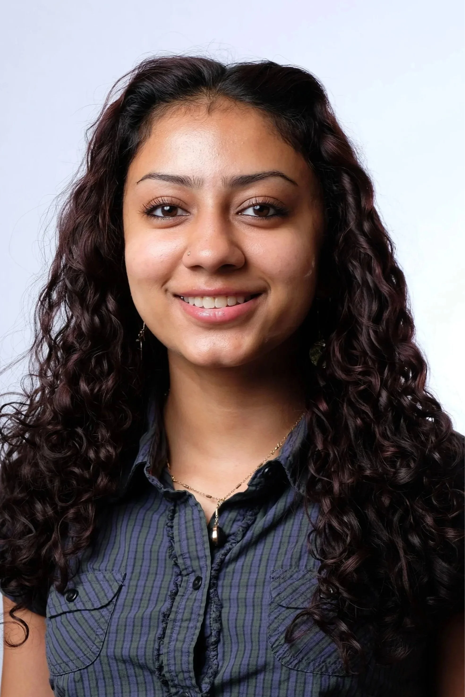 Tara Sawhney - A woman with long, curly dark hair smiling at the camera, wearing a dark striped shirt and a gold necklace with a pendant.