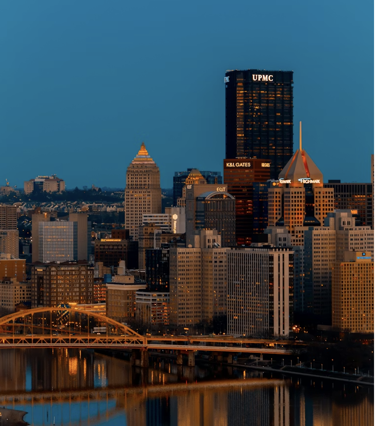 Night view of downtown Pittsburgh with illuminated buildings, bridges over the river, and city lights reflecting on the water.