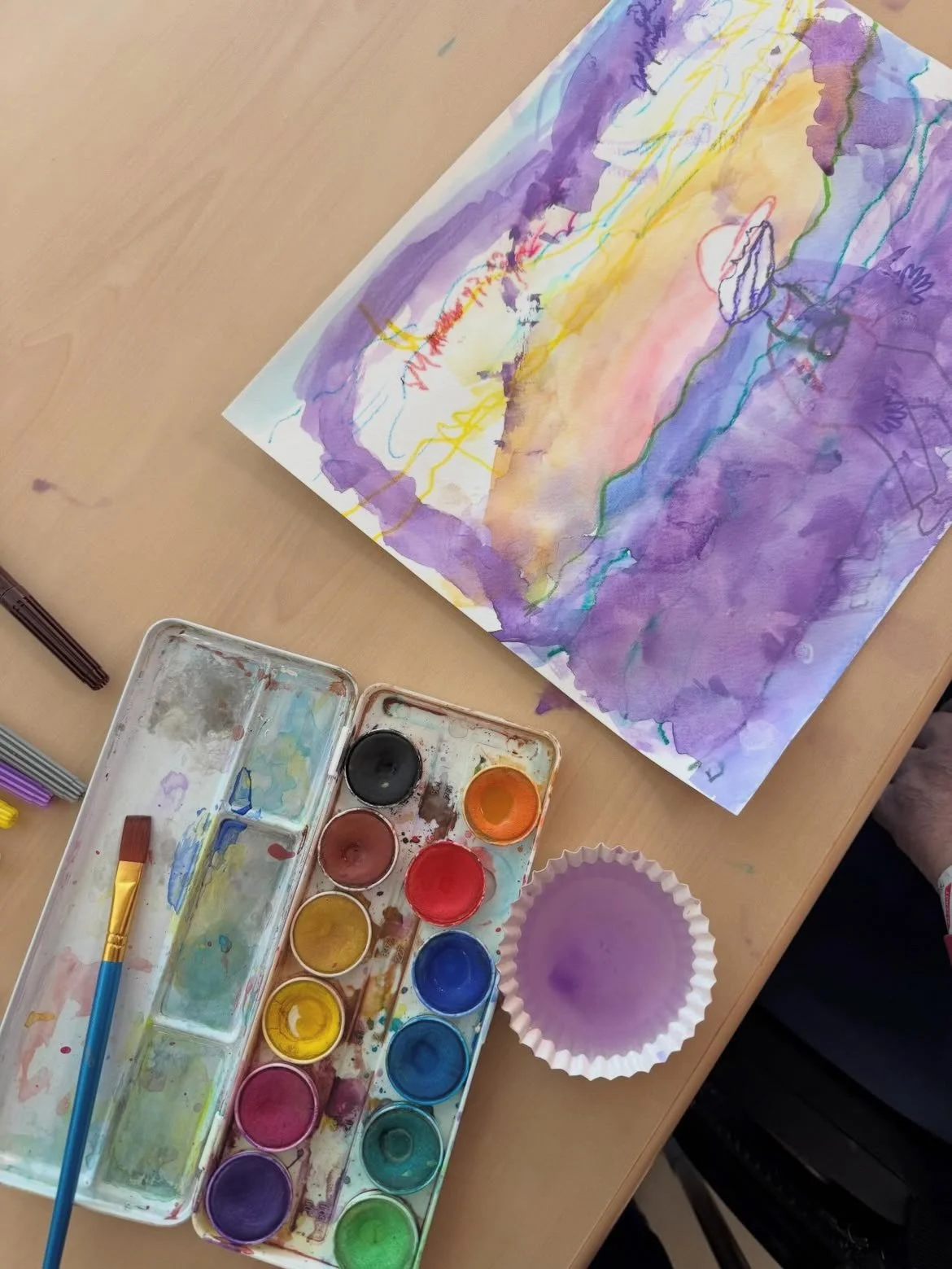 Watercolor painting on paper in progress with a paint palette, brushes, and a purple cupcake liner containing purple paint on a light wooden table.