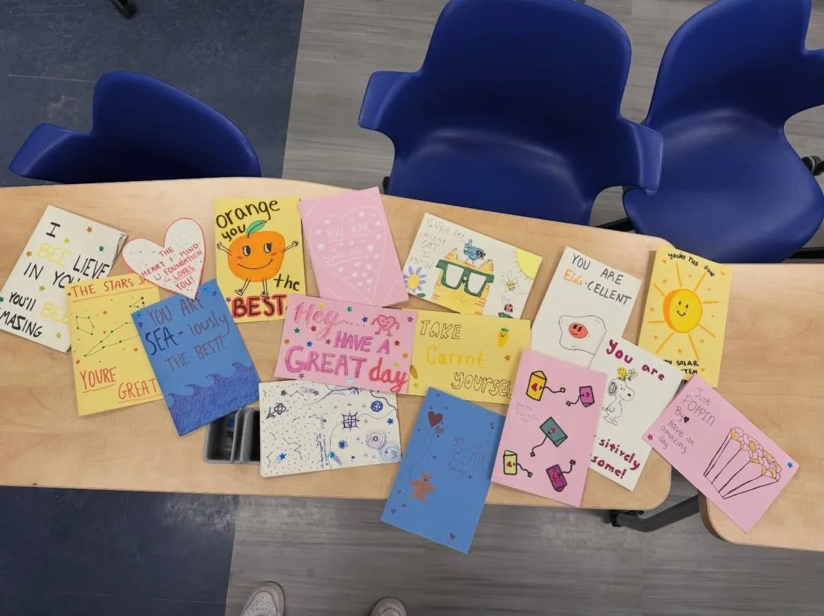 Collection of colorful handwritten and illustrated greeting cards and notes on a wooden table, with blue chairs around, in a classroom or community setting.