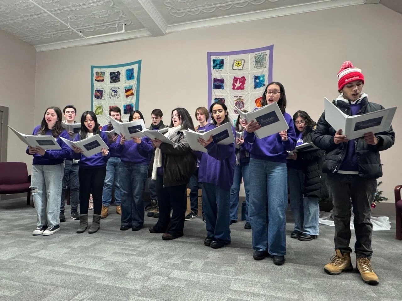 A group of young people singing in a room, holding sheet music, some wearing purple hoodies. The room has decorative quilts hanging on the wall and a small Christmas tree on the side.