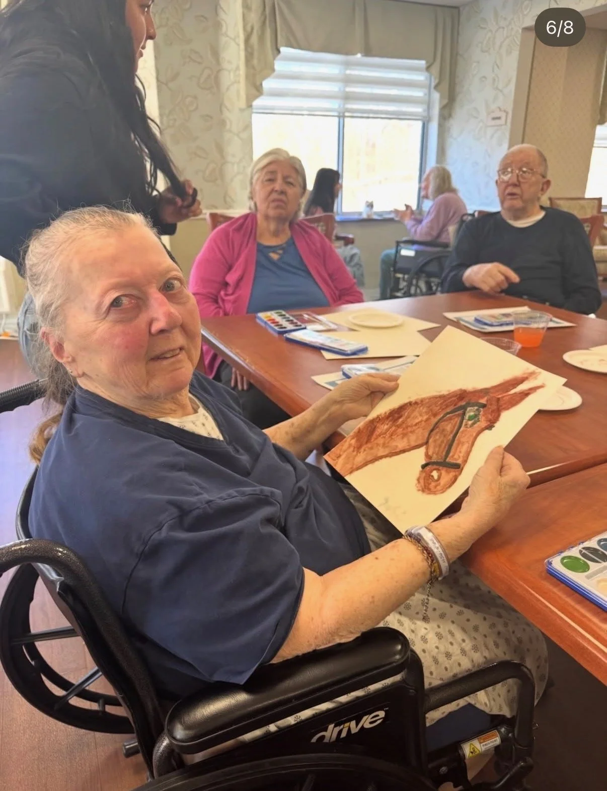 An elderly woman in a wheelchair holding a painting of a brown horse, sitting at a table in a dining area with other seniors, some with cameras, with light coming through windows.