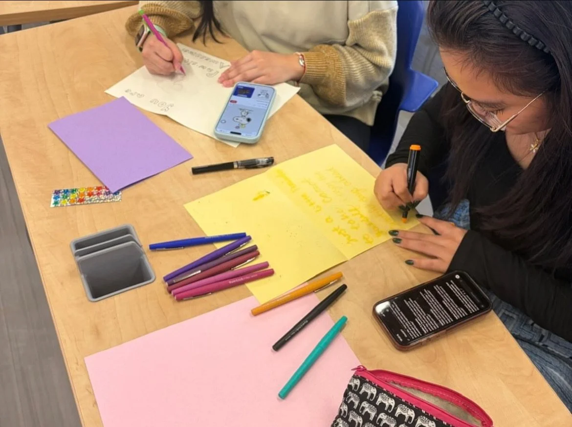 Two women sitting at a wooden table working on colorful posters with markers, pens, and paper. One woman is writing on a yellow poster while the other is drawing and holding a pink pen. There are various art supplies and a smartphone on the table.