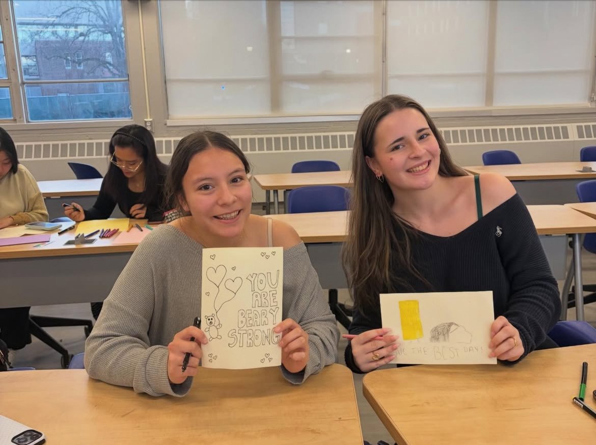 Two girls sitting at a table in a classroom, smiling and holding handmade cards, with other students working in the background.