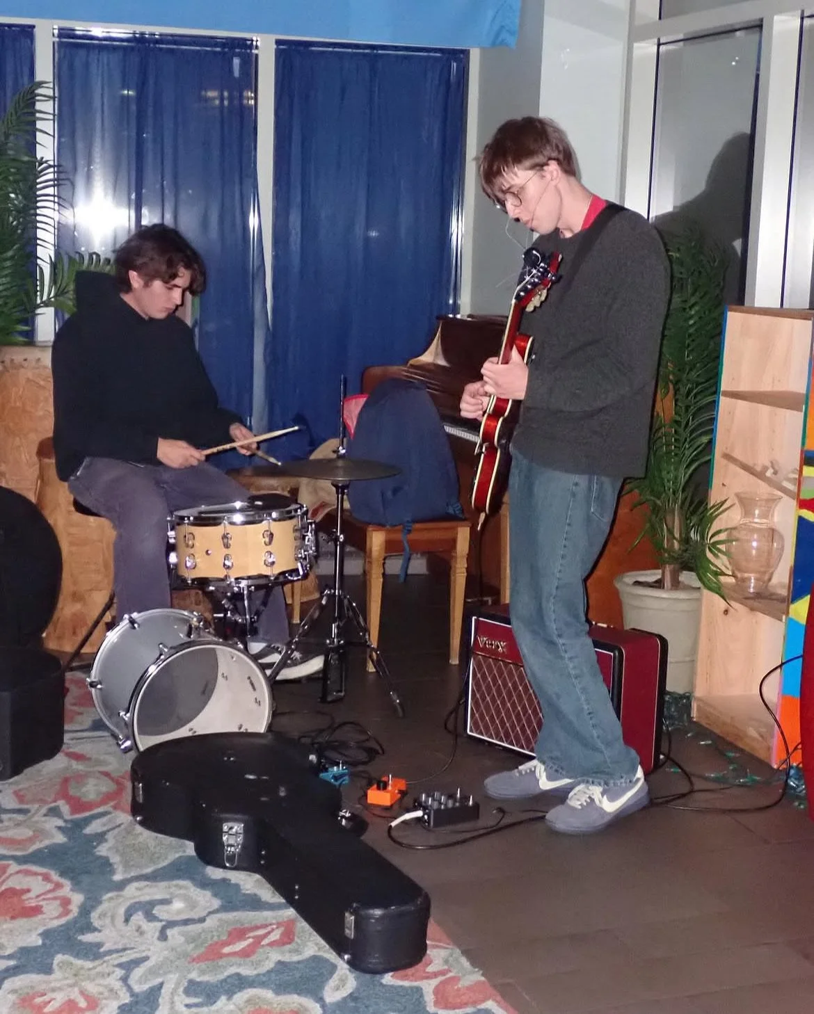 Two young men playing musical instruments in an indoor setting. One is on drums, and the other is playing an electric guitar. There is music equipment on the floor and a black guitar case. The background features blue curtains, plants, and wooden she