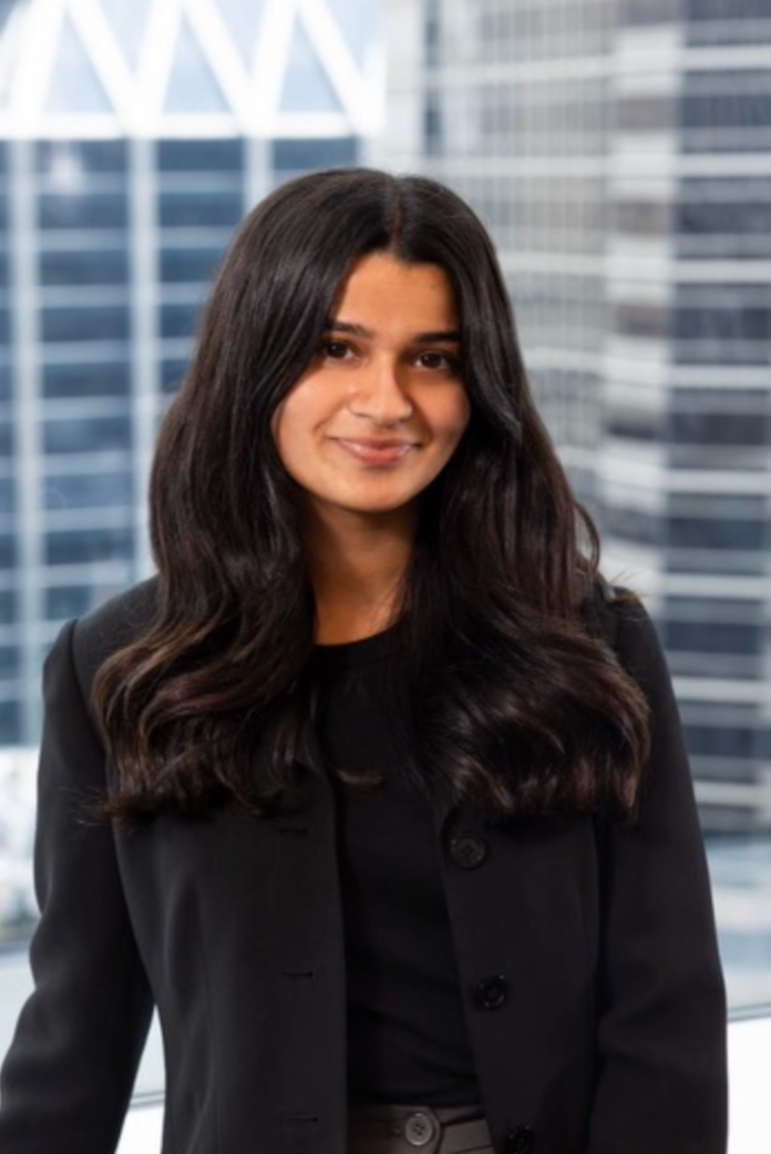 Aliza Vriji - A woman with long dark hair smiling, wearing a black blazer, standing in front of tall glass office buildings.