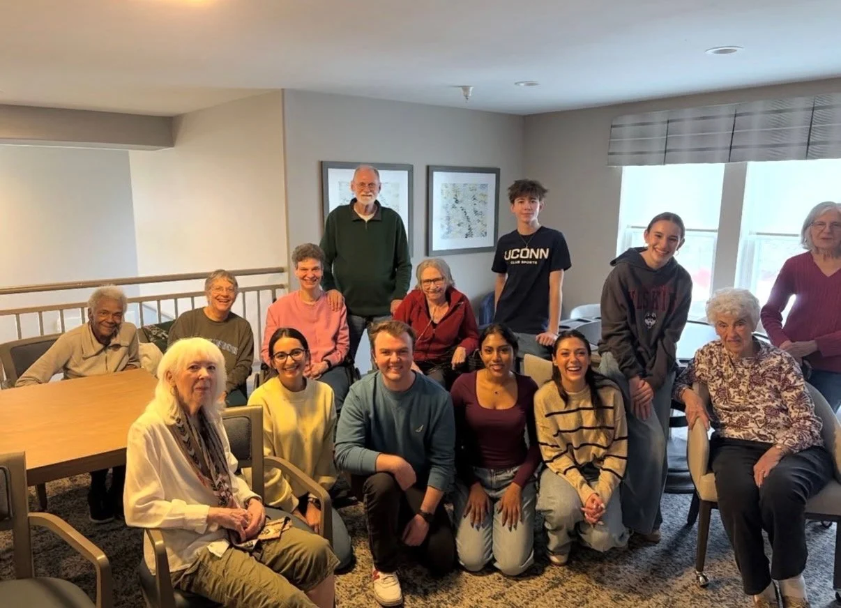 Group of fifteen people, mostly seniors, gathered in a room with large windows, seated and standing for a photo, some smiling and some looking serious.