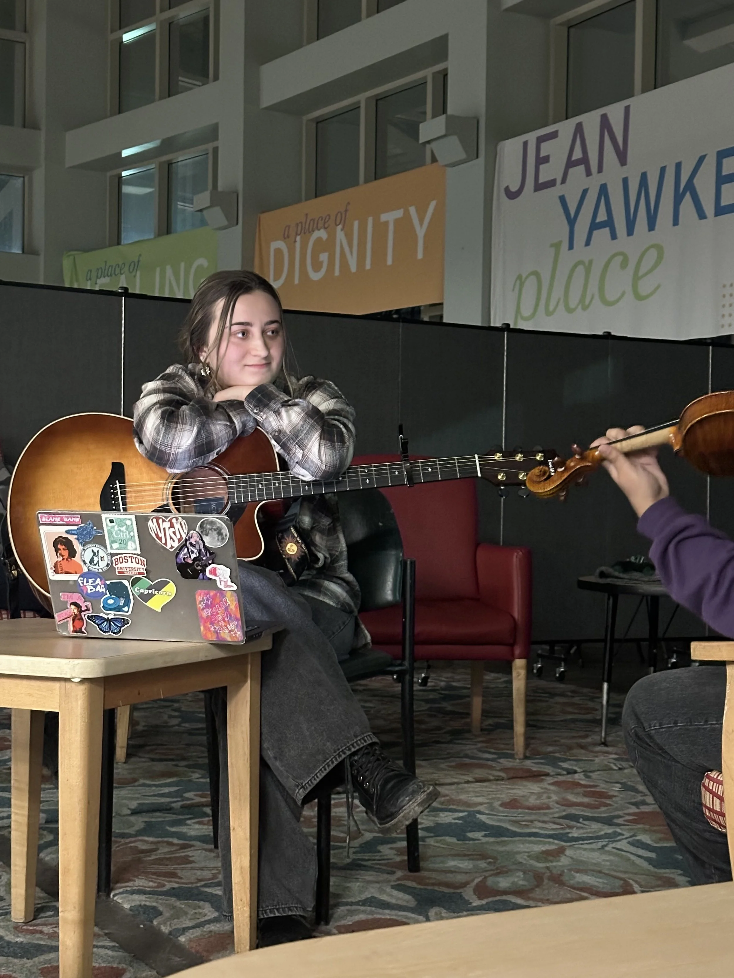 A young woman sitting on a chair with a guitar on her lap, listening to someone playing a violin. Her face shows a faint smile and she is resting her chin on her crossed arms. The setting appears to be indoors, possibly a community center or a room f