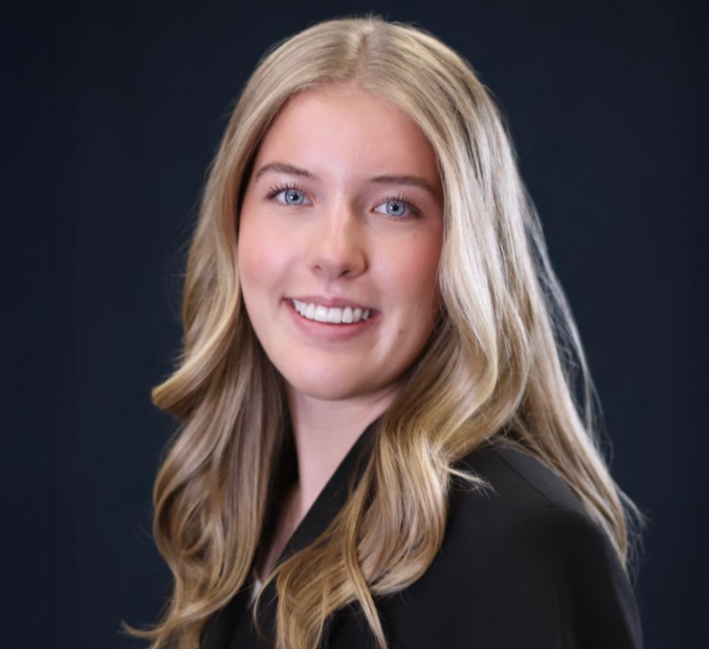 A young woman with long, blonde hair and blue eyes, smiling, dressed in a black blazer, against a dark background.