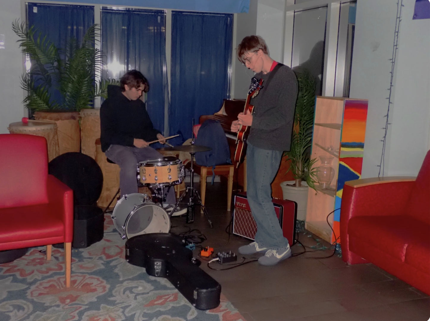 Two young men playing musical instruments in a cozy indoor room. One is sitting on a chair with drumsticks, playing a small drum set. The other is standing, playing an electric guitar. The room has red couches, a patterned rug, a bookshelf, potted plants, a colorful painting, and a blue curtain.