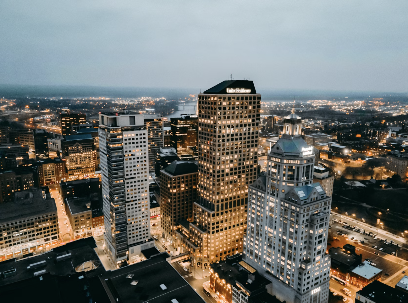 An aerial view of a cityscape at dusk, featuring tall skyscrapers, illuminated streets, and a historic white clock tower surrounded by modern buildings.