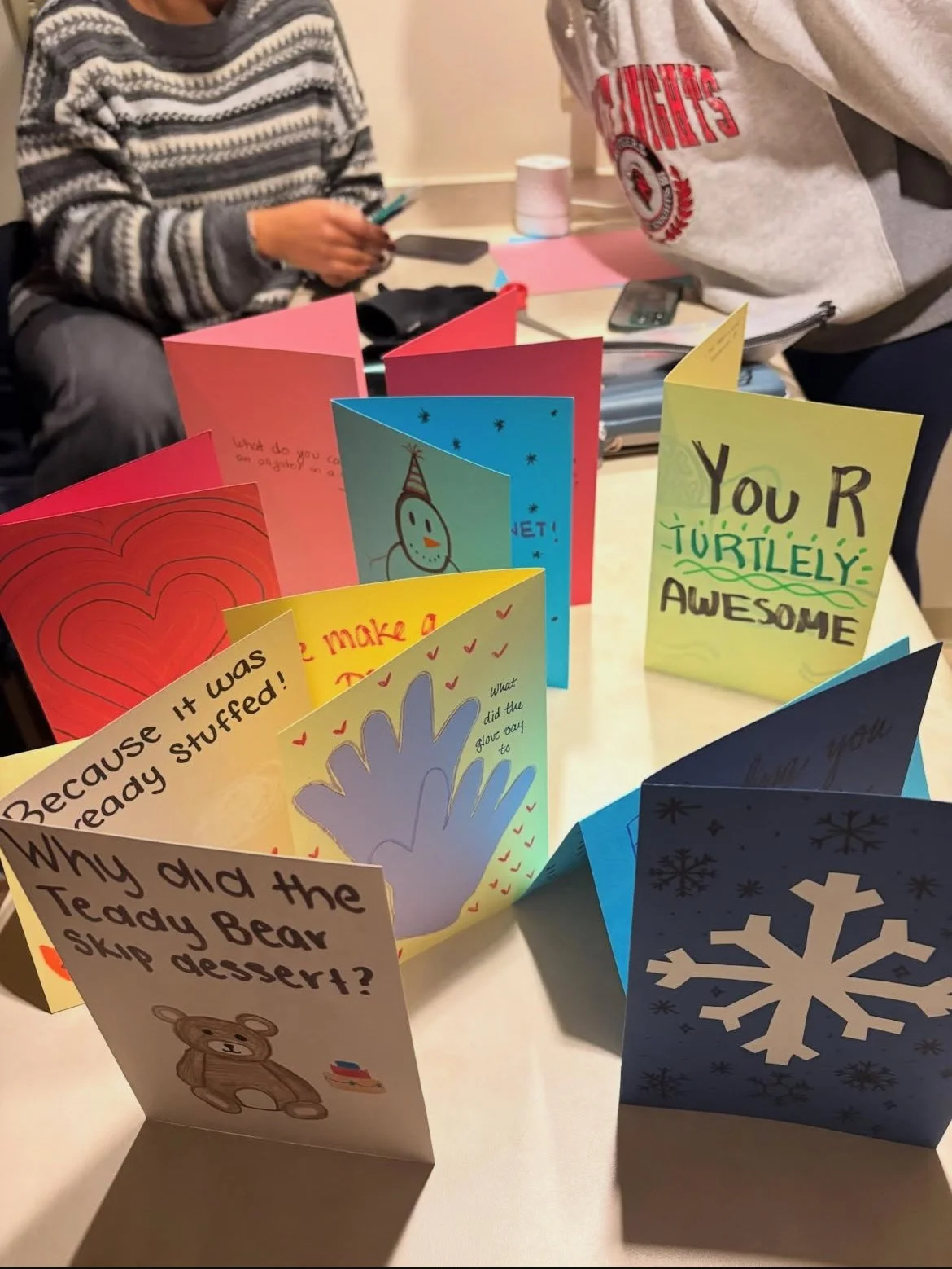 Colorful folded greeting cards on a table with handwritten and printed messages, including a snowflake, a teddy bear, and a handprint, with people sitting in the background.