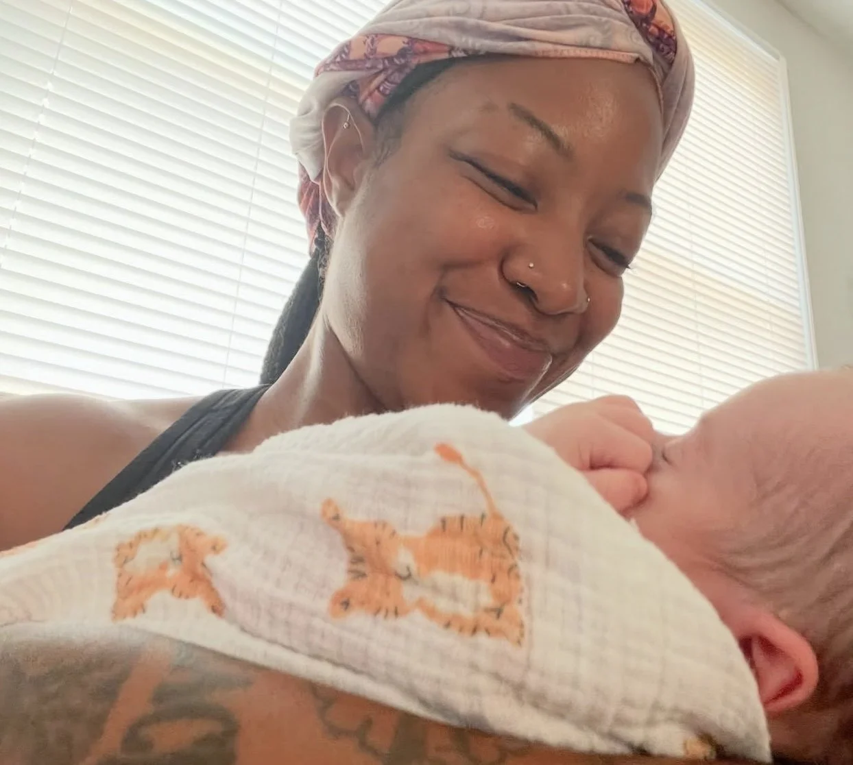 A non-binary person with a pink headwrap is smiling at a newborn baby wrapped in a blanket with tiger patterns.