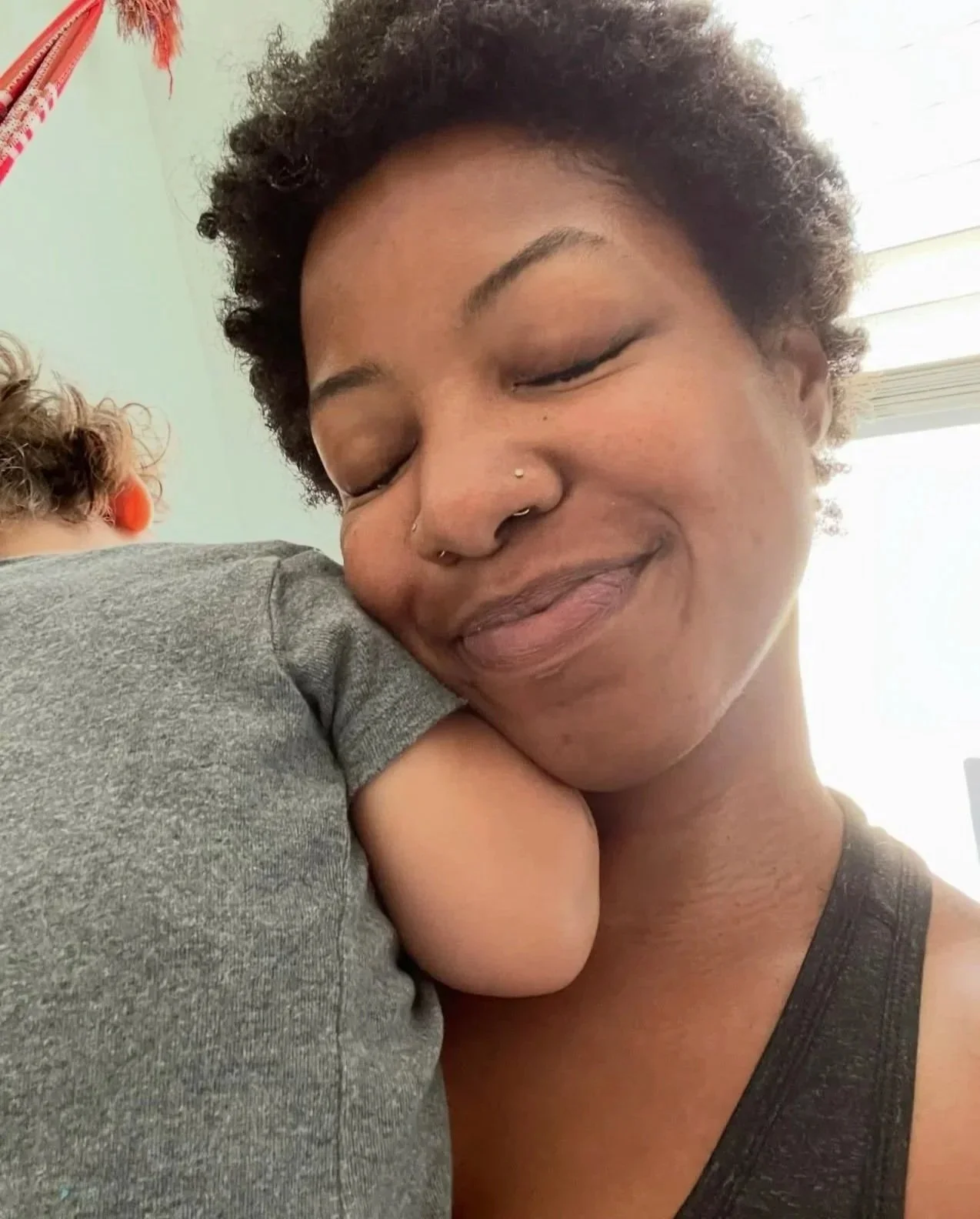 A non-binary person with short curly hair, wearing a dark tank top, hugging a child with curly hair and a gray shirt, her eyes closed and smiling softly.