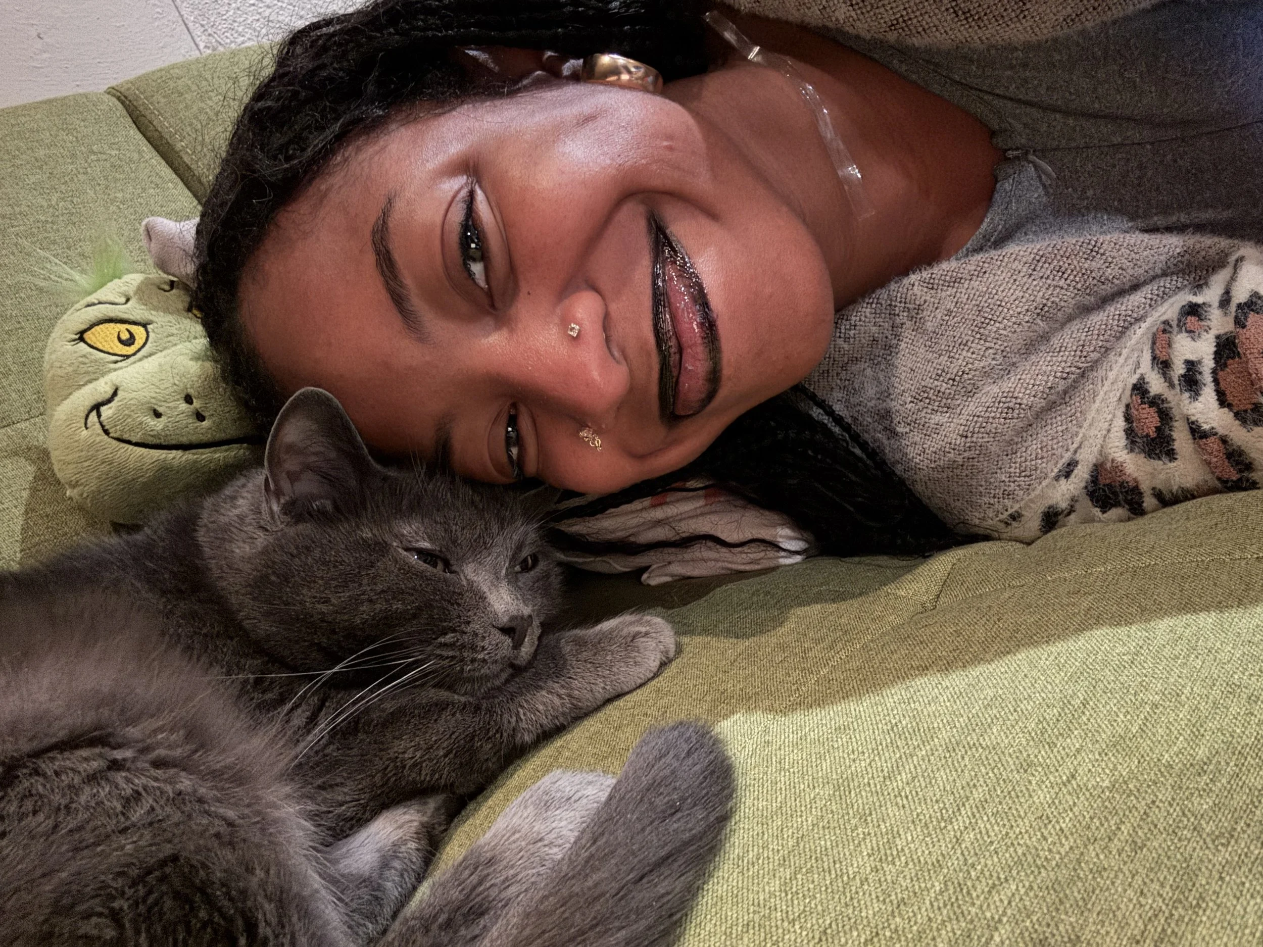 A non-binary person with dark hair and jewelry lying on a green couch with a gray cat resting its head on her chest, both appearing relaxed.