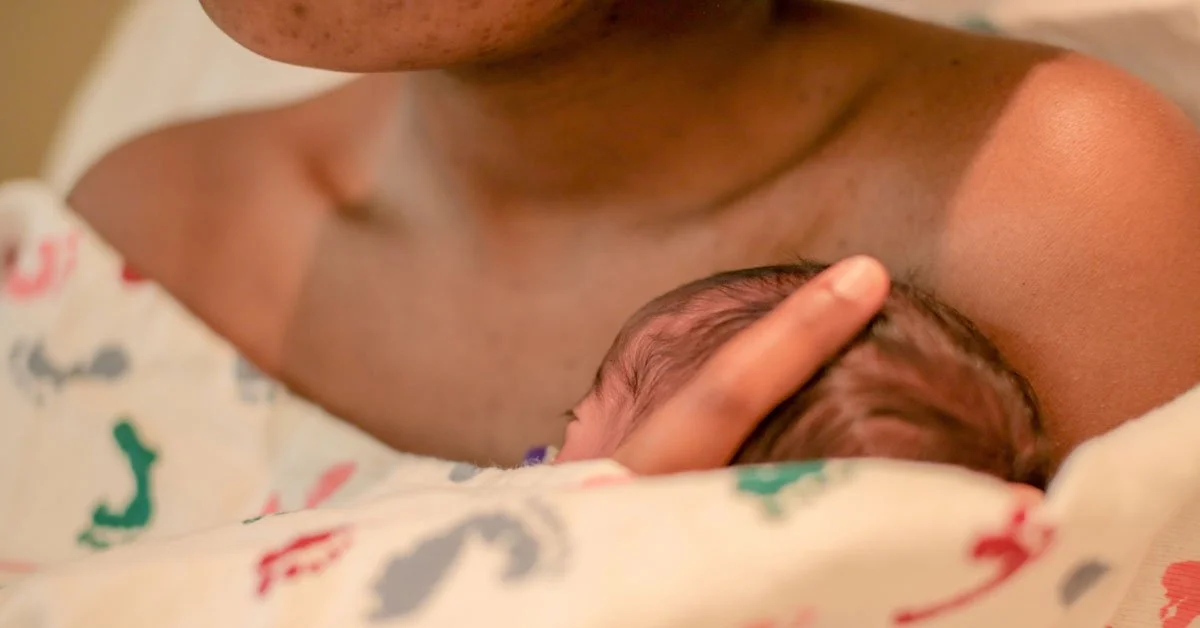 Close-up of an adult cradling a newborn baby against their chest, with the baby's head resting in the adult's hand, wrapped in a patterned blanket.