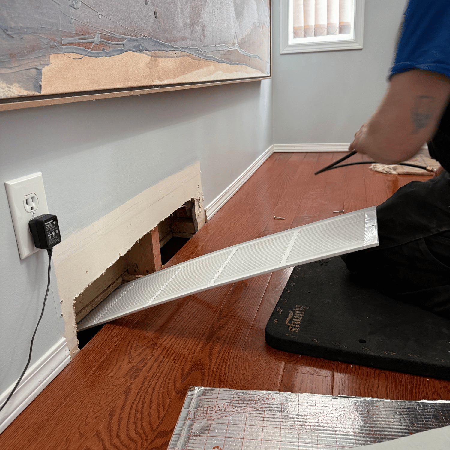 A person installing or removing a vent cover on a wall with hardwood flooring, next to an electrical outlet.