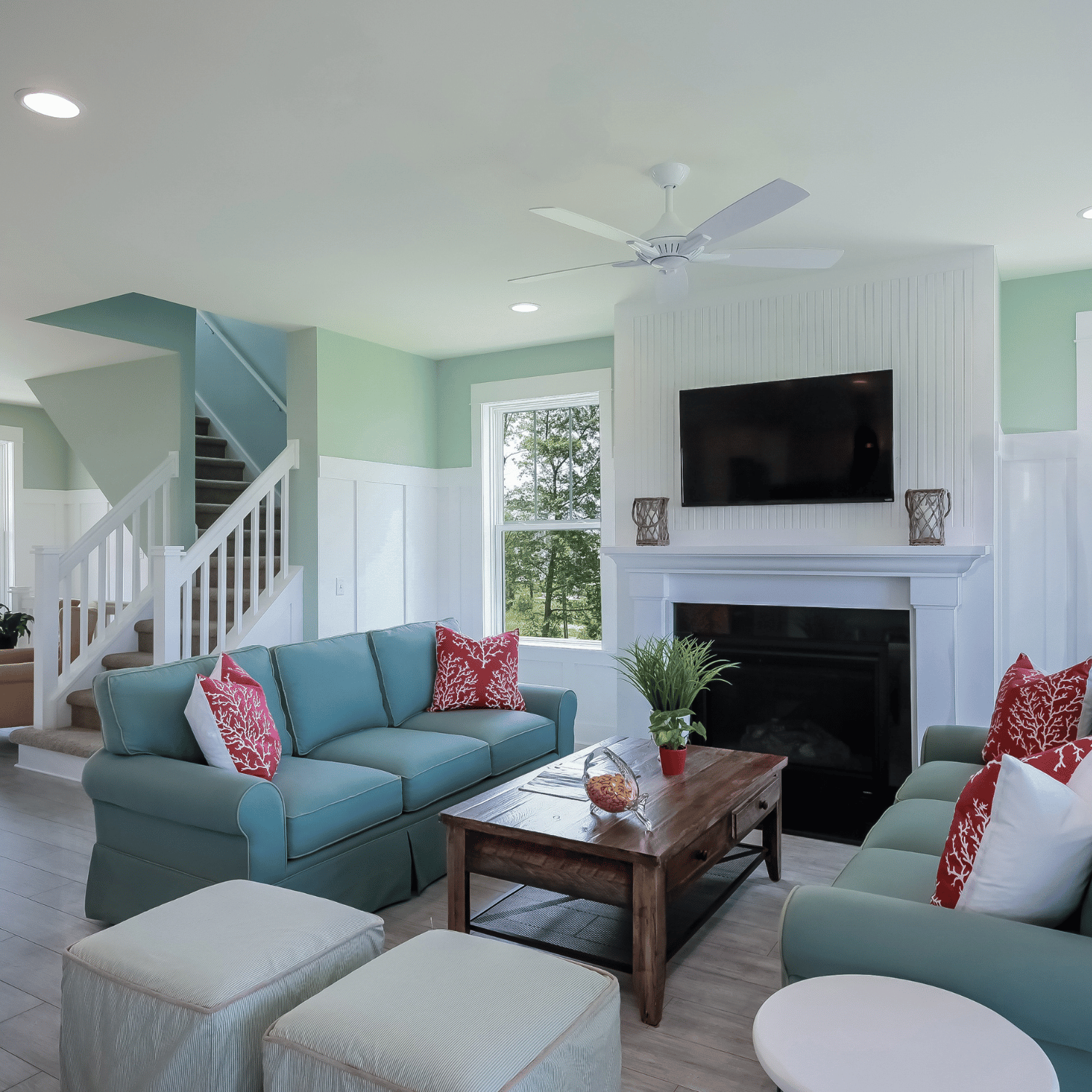 Bright living room with teal sofas, pillows, a wooden coffee table, two ottomans, a fireplace, a flat-screen TV, staircase, and large window with green wall accents.