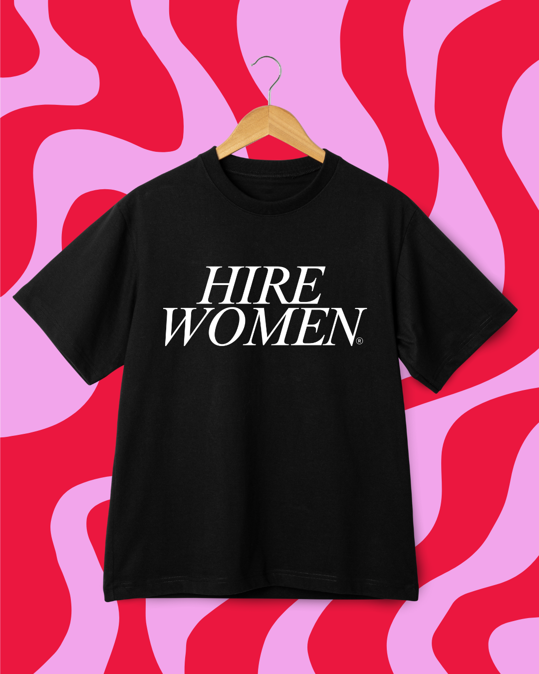 Black T-shirt with white text that says 'HIRE WOMEN' hanging on a wooden hanger against a pink and red abstract background.