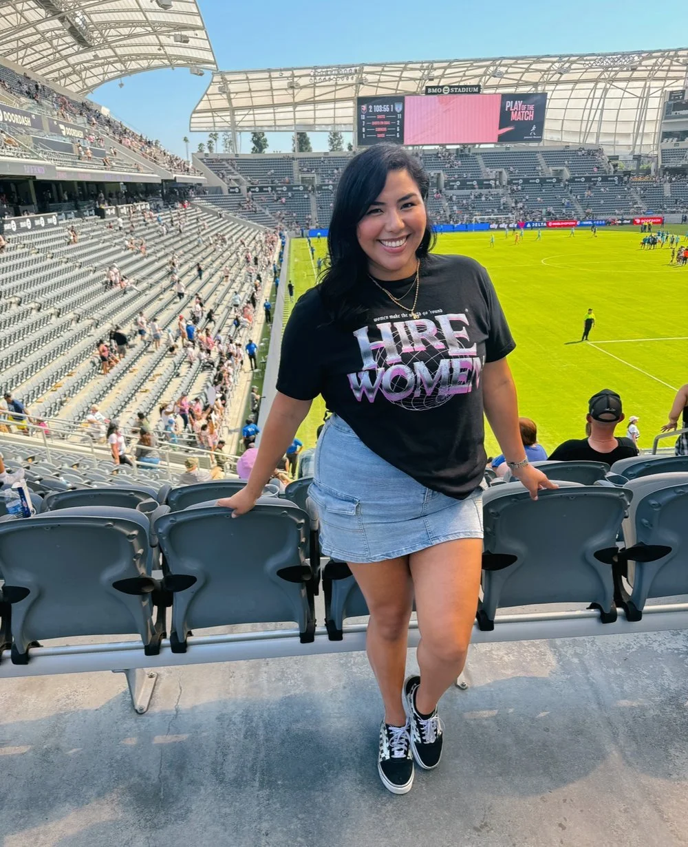 A person in a stadium wearing a black 'Hire Women' T-shirt and denim skirt, smiling and posing for a photo. The stadium has empty gray seats and a green soccer field with other people and players in the background.