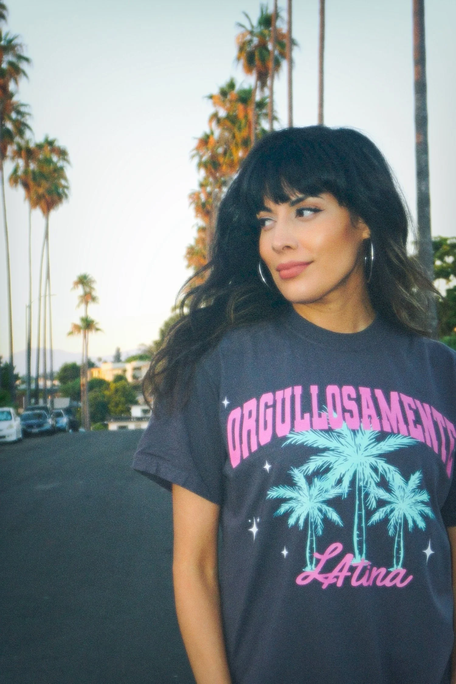 A woman wearing a "Orgullosamente Latina" T-shirt, standing on a street lined with palm trees.