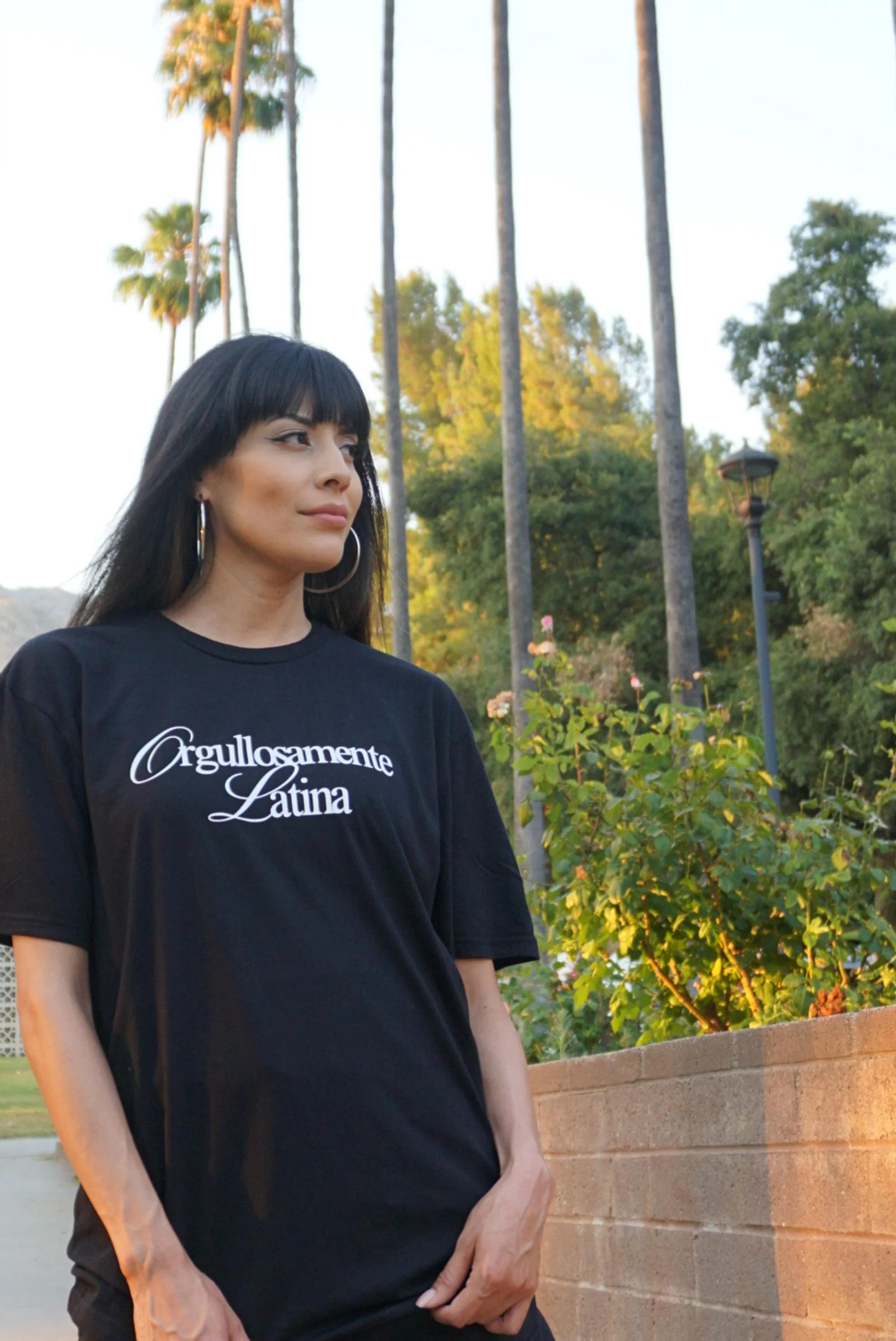Woman with black hair and hoop earrings standing outdoors near a brick wall, wearing a black T-shirt with white text that says "Orgullosamente Latina," and palm trees in the background during late afternoon or early evening.
